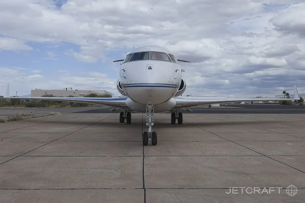 A head-on view of a white Bombardier Global 6000 business jet with blue and grey stripes, parked on an airfield.