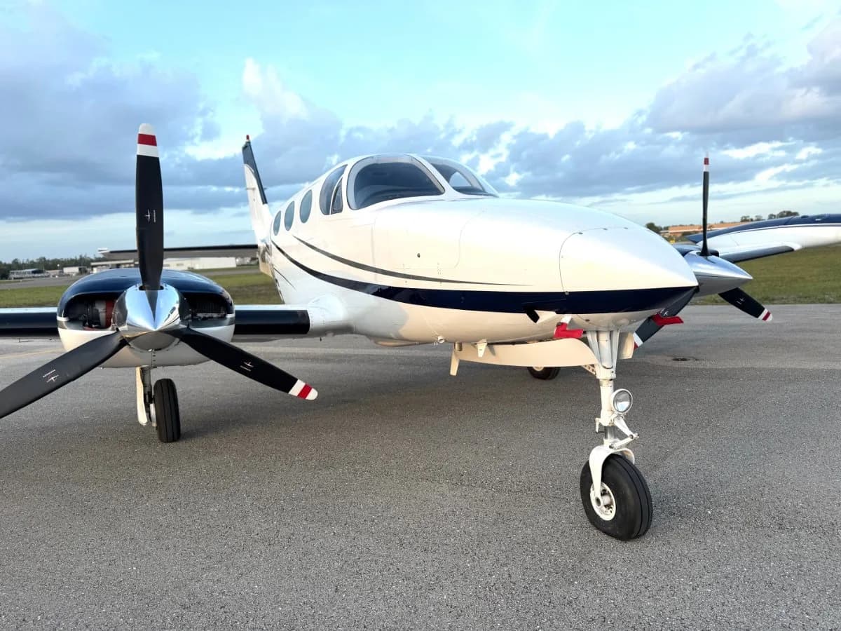 A white twin-engine propeller aircraft with a dark blue stripe is parked on an airport tarmac under a cloudy sky.