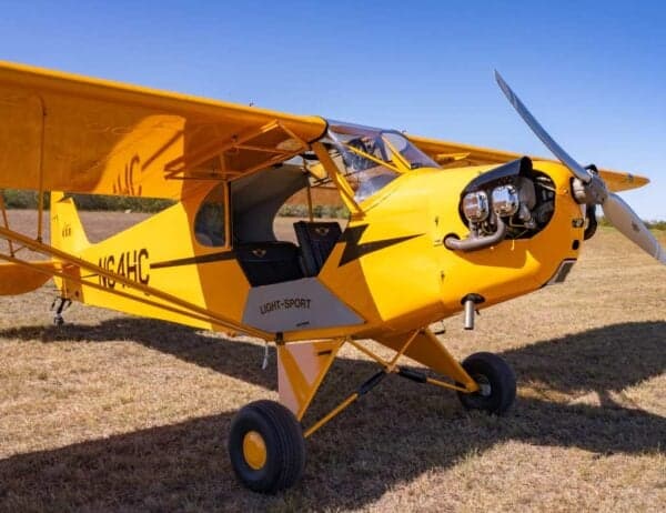A front-side view of the yellow Legend Cub aircraft, registration N64HC, with its engine exposed, parked on a grassy field.