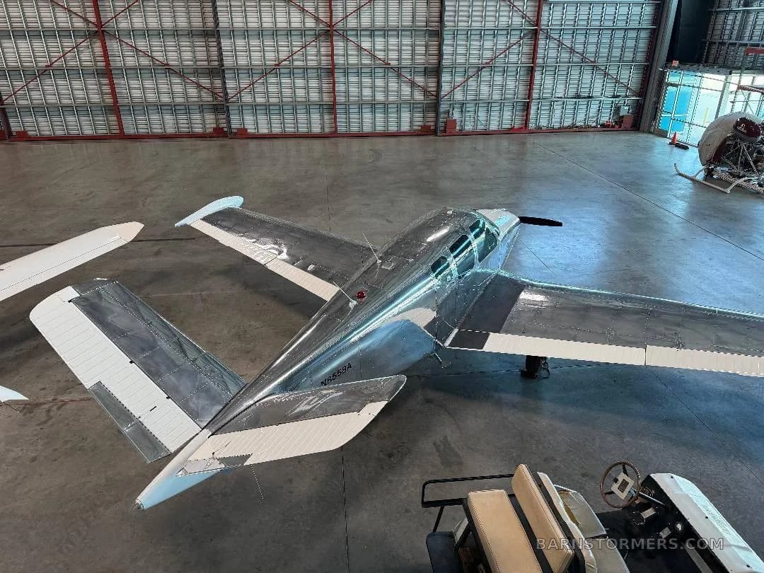 An overhead view of a bare metal, V-tail aircraft (N8558A) with white wingtips and tail surfaces, parked in a hangar.
