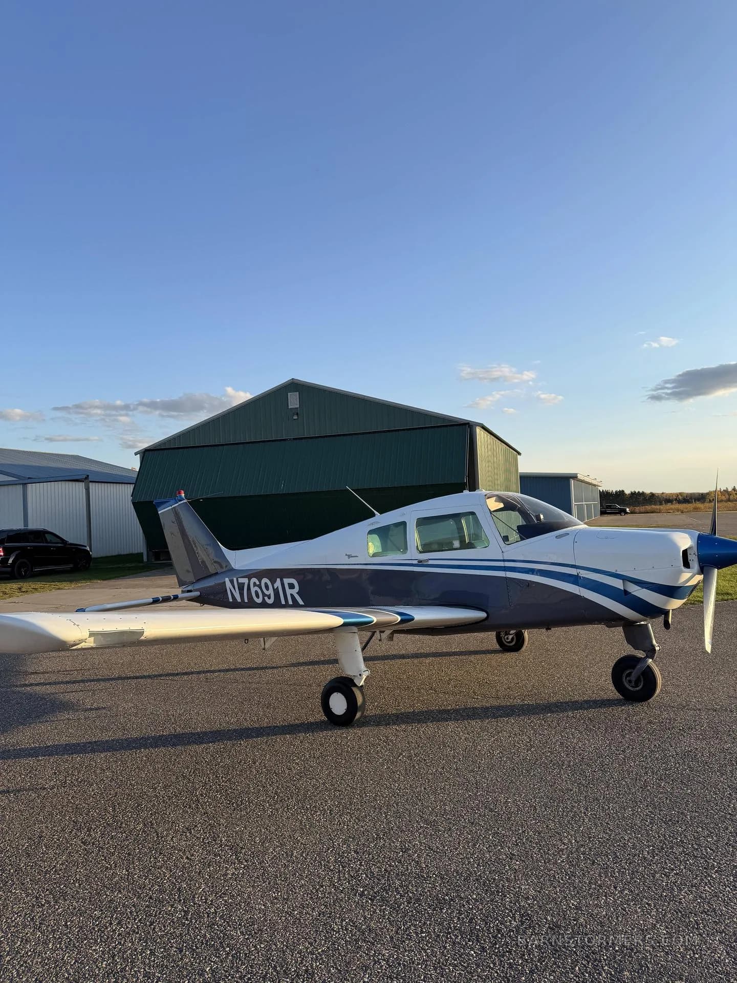 A white and blue single-engine aircraft (N7691R) parked on an asphalt ramp in front of a green hangar under a blue sky.