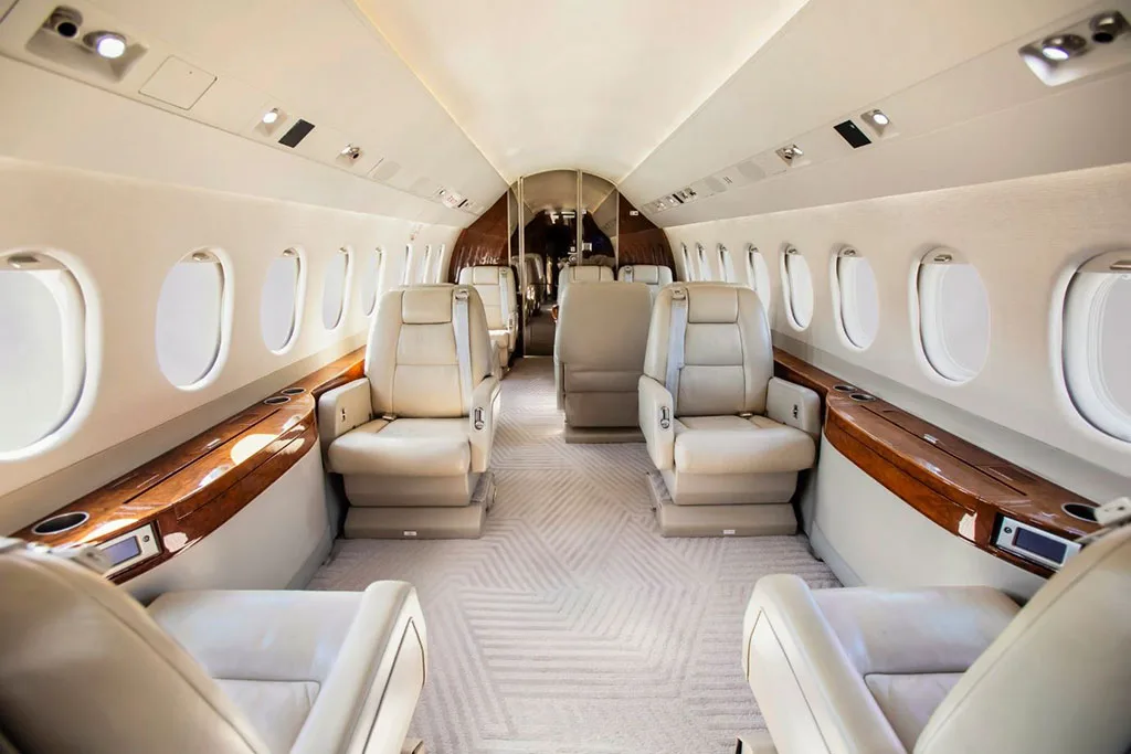 A view down the aisle of a private jet's cabin, showing multiple rows of light-colored leather seats and polished wood accents.