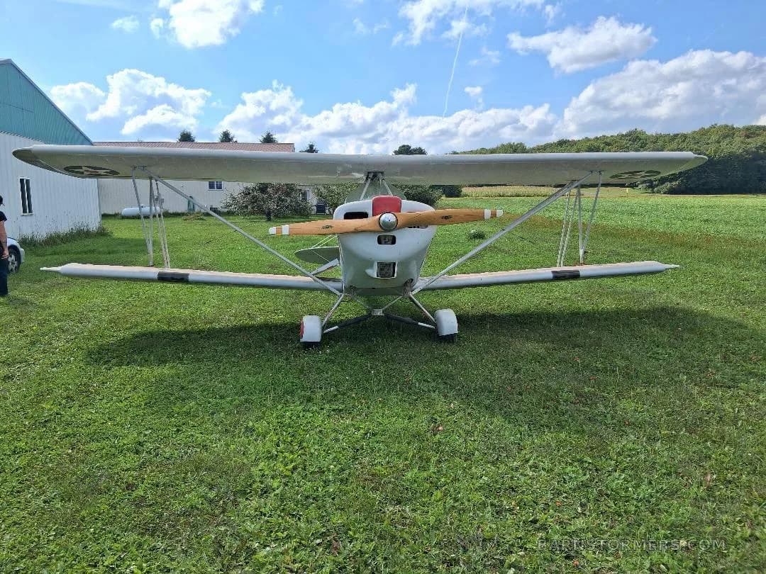 A white biplane with a red nose cone and wooden propeller, viewed from the front, parked on green grass under a blue sky.