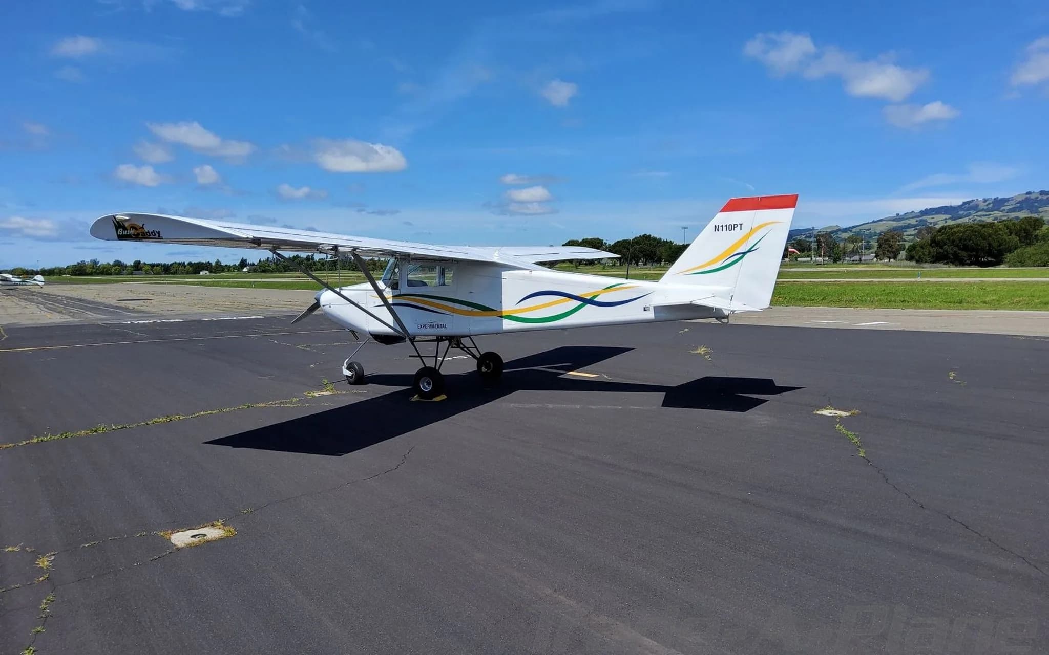 A white BushCaddy aircraft with green, yellow, and black wavy stripes, parked on a sunny tarmac with hills in the distance.