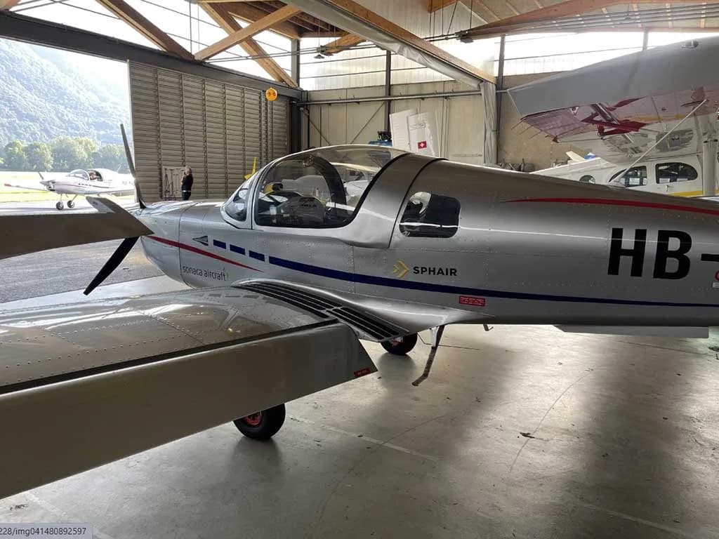 A silver Sonaca aircraft with registration HB-KGF parked in a hangar, viewed from the front-left side.