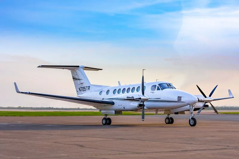 A white Beechcraft King Air 350i with tail number N725TR is parked on an airfield under a blue sky.