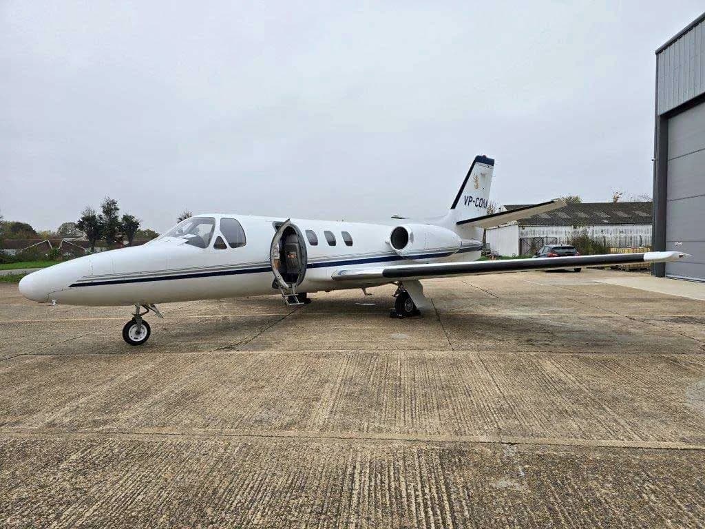 A white Cessna Citation jet with blue and grey stripes, registration VP-COM, parked on a tarmac with its main door open.