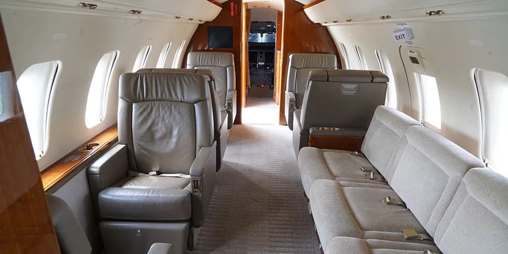 The interior cabin of a private jet features light grey leather seats, a beige sofa, and wooden accents, looking forward towards the cockpit.