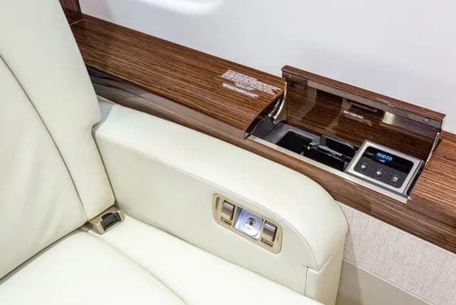 A close-up of a light leather aircraft seat armrest with controls and a side panel featuring a hidden compartment with audio controls.