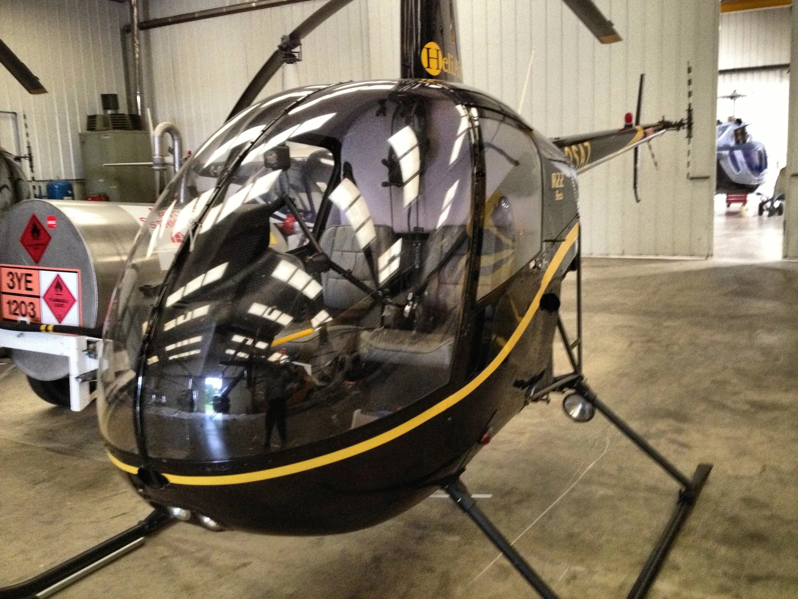A black Robinson R22 helicopter with a yellow stripe parked inside a hangar, reflecting the overhead lights.