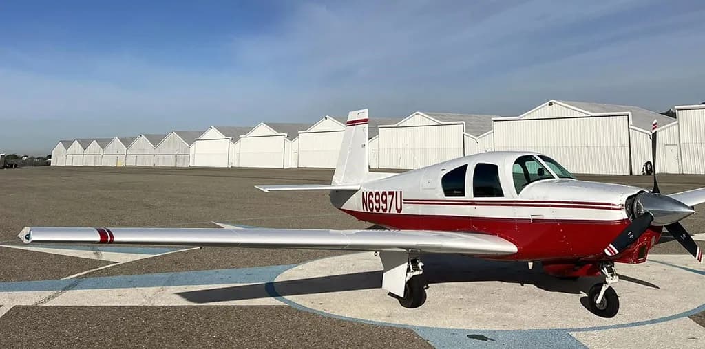 A white and red single-engine aircraft, N6997U, with a propeller, parked on an airfield in front of a row of hangars.