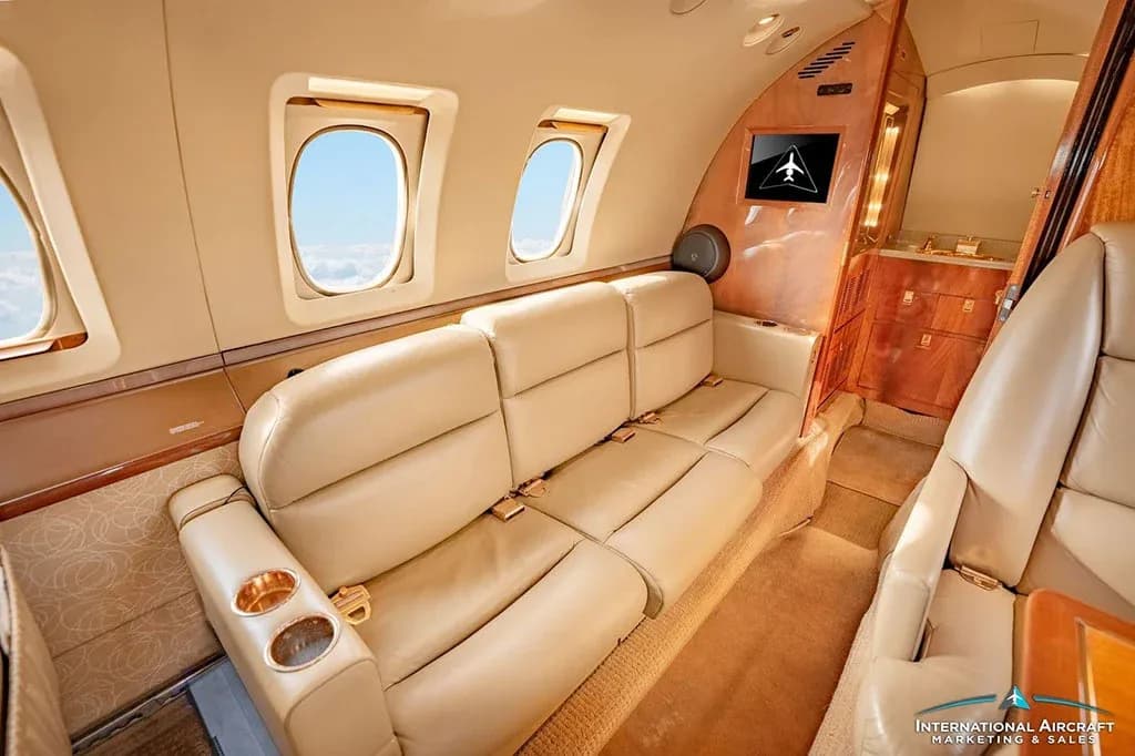 A comfortable beige leather couch-style seat is visible in the private jet's cabin, next to a wooden cabinet.