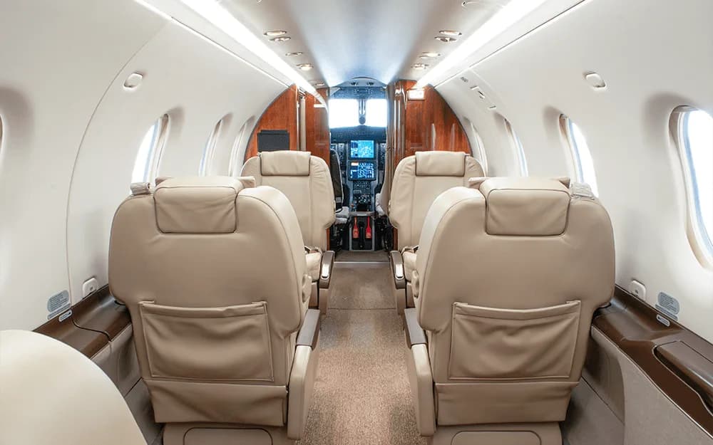 Interior view of a private aircraft cabin with beige leather seats, looking forward towards the cockpit.