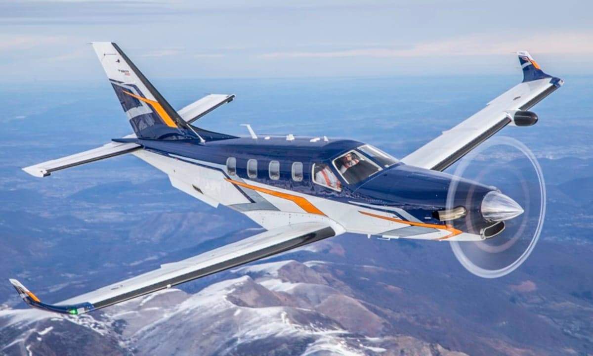 A blue, white, and orange TBM 980 turboprop aircraft flying over snowy mountains.