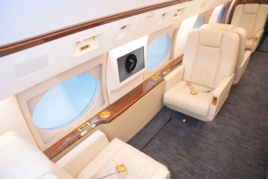 A view of the private jet cabin showing cream leather seats, large oval windows, and polished wood trim.