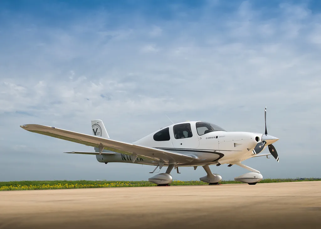 A white and silver Cirrus SR22T aircraft, N171TX, parked on a tarmac under a partly cloudy blue sky with a green field in the background.