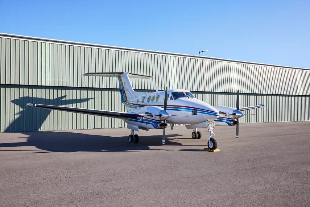 Side view of a Beechcraft King Air with a white and blue paint scheme parked in front of a hangar.