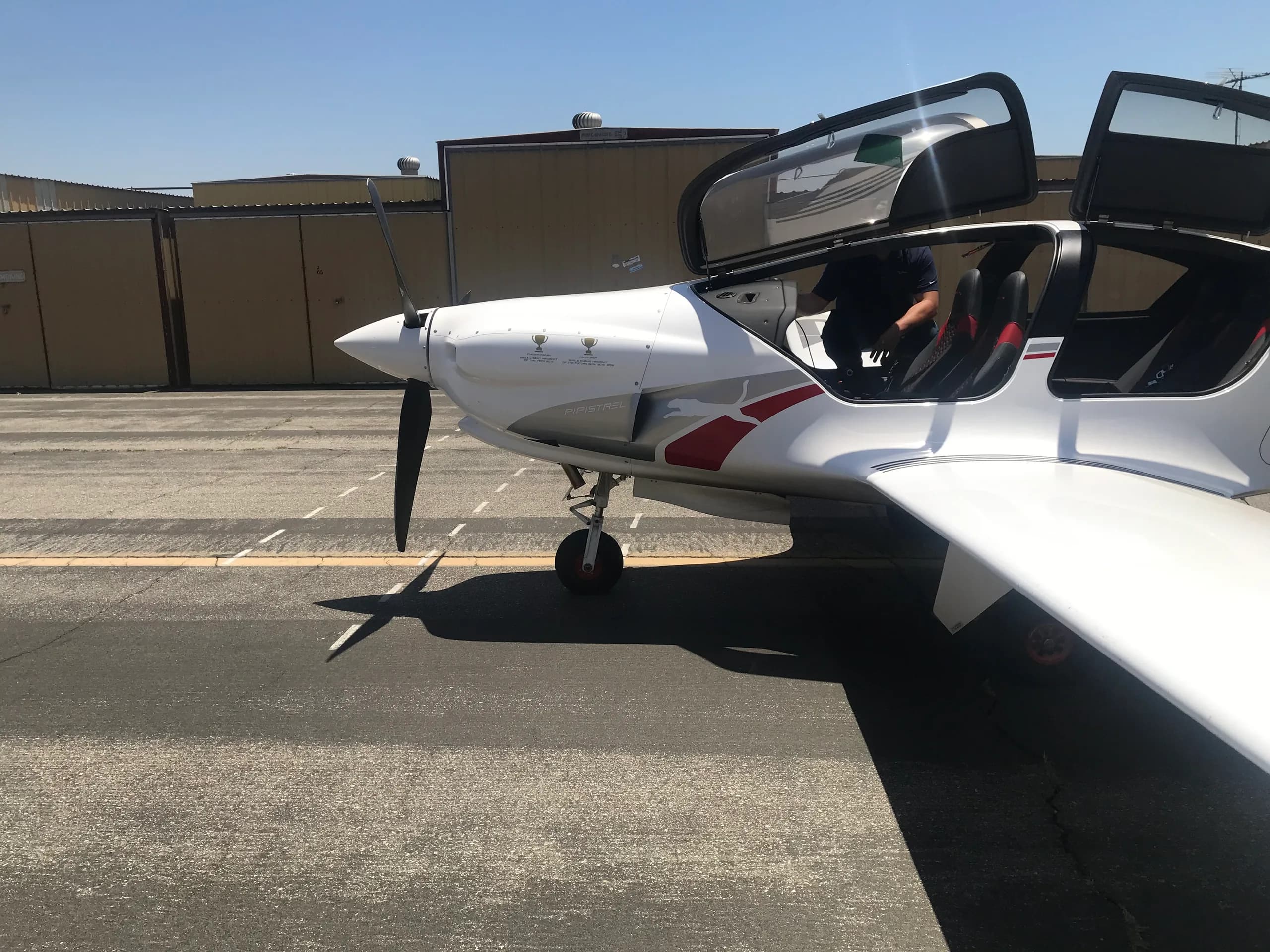 A white Pipistrel Panthera aircraft with red and gray accents, parked on an airfield with its canopy open and a person in the cockpit.