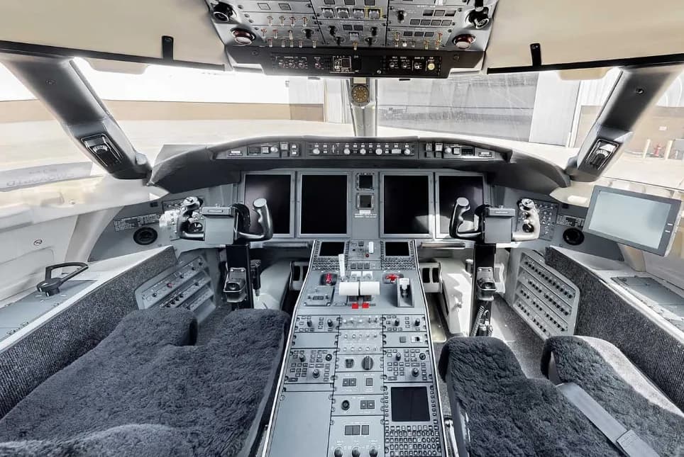 The spacious cockpit of a modern business jet features multiple large display screens, a central control panel, and dark sheepskin seat covers.