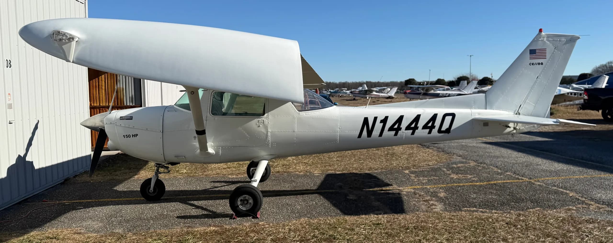 A full side view of the white Cessna 150 aircraft, registration N1444Q, parked on an airfield with other aircraft in the distance.