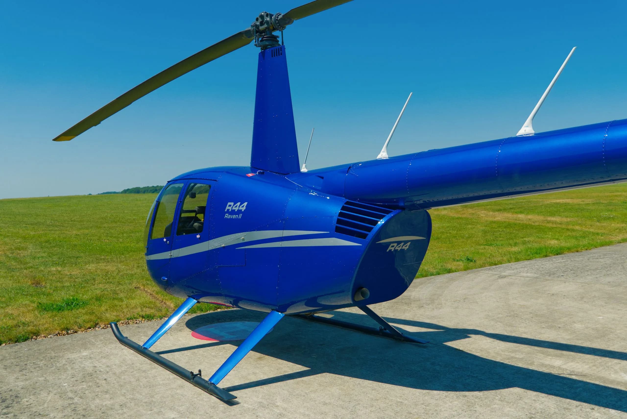 A blue Robinson R44 Raven II helicopter is parked on a concrete pad with grass in the background.