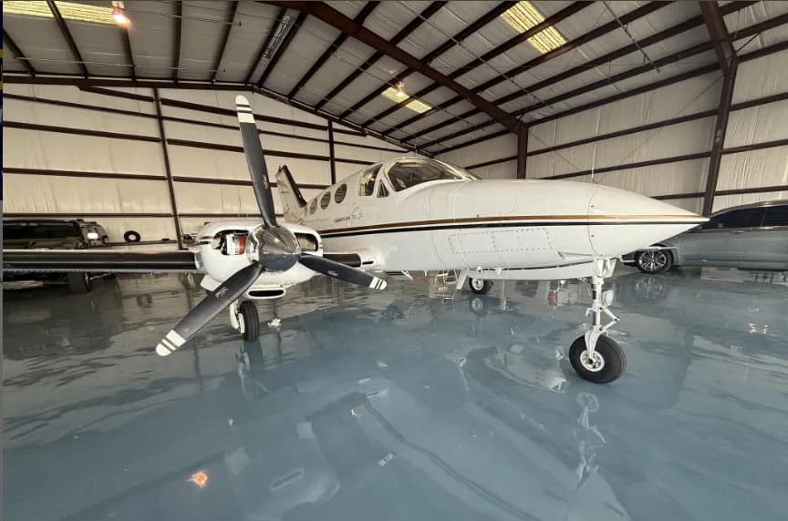 A white Cessna 414 aircraft with gold and black stripes, parked in a hangar, viewed from the front-left.