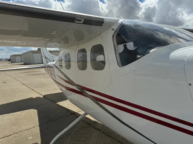 A close-up side view of the cockpit and cabin windows of a white Cessna 206 with red and grey stripes.