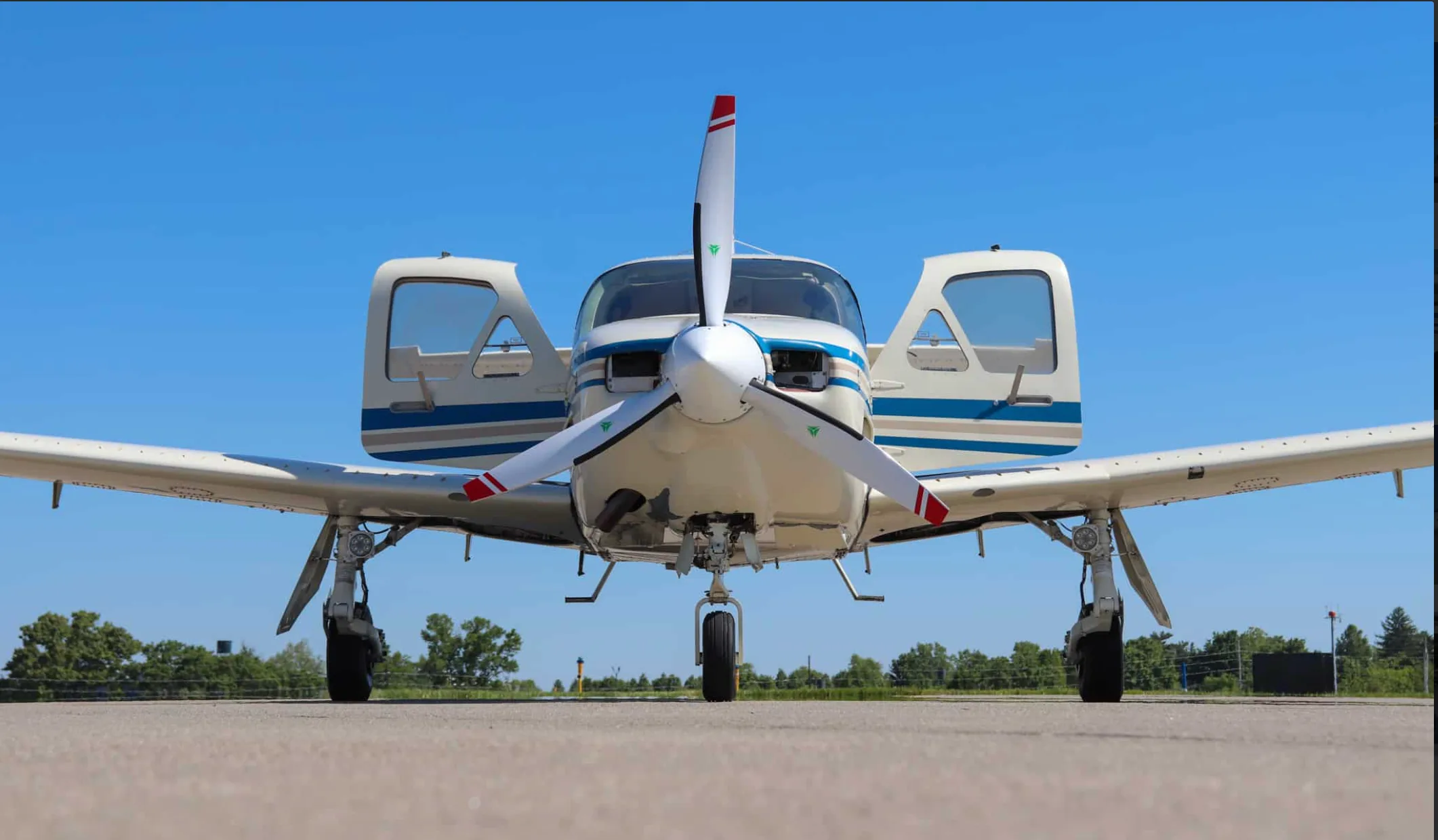 Low-angle front view of a white single-engine propeller aircraft with blue and tan stripes, parked on an airfield with doors open.
