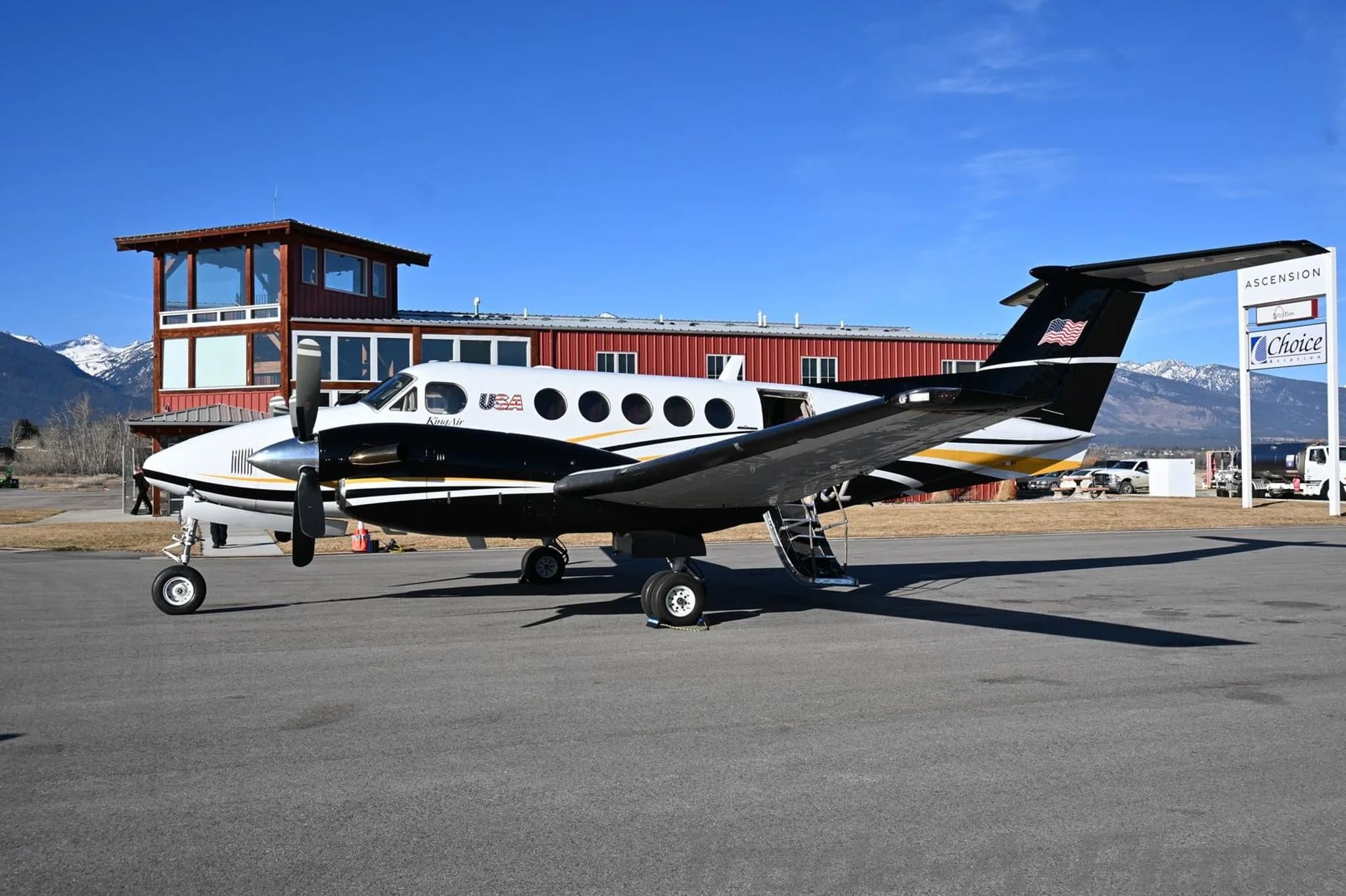 A white and black Beechcraft King Air 200 with gold stripes parked on an airfield, viewed from the rear-left.