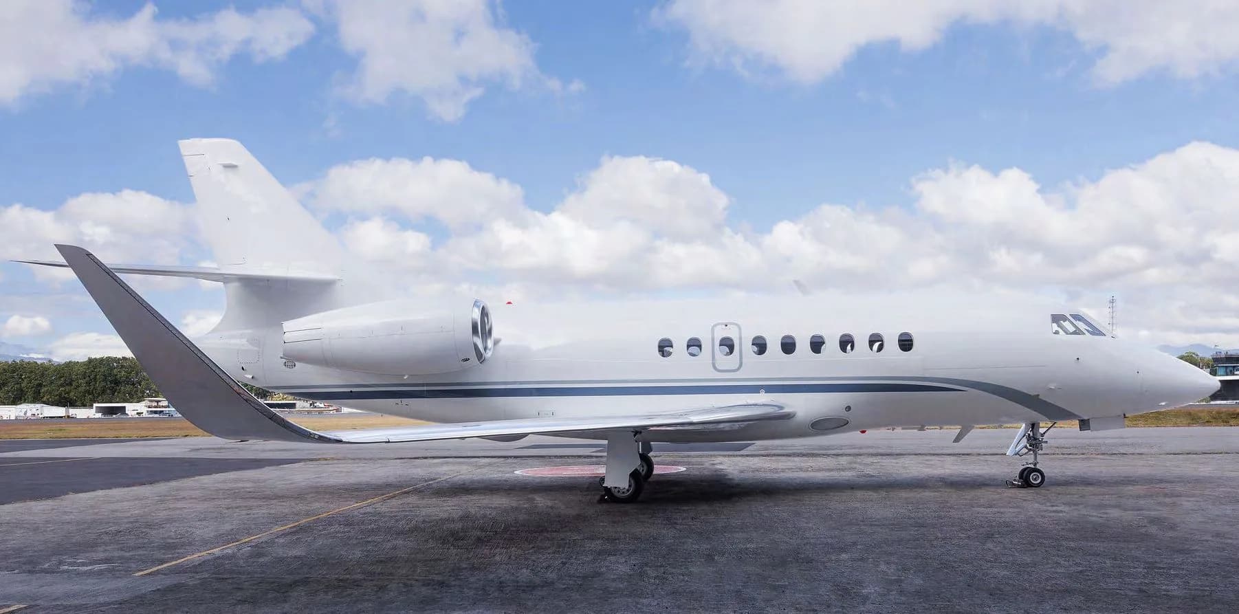 A white Dassault Falcon 2000LXS business jet with blue and grey stripes parked on an airport tarmac under a cloudy sky.