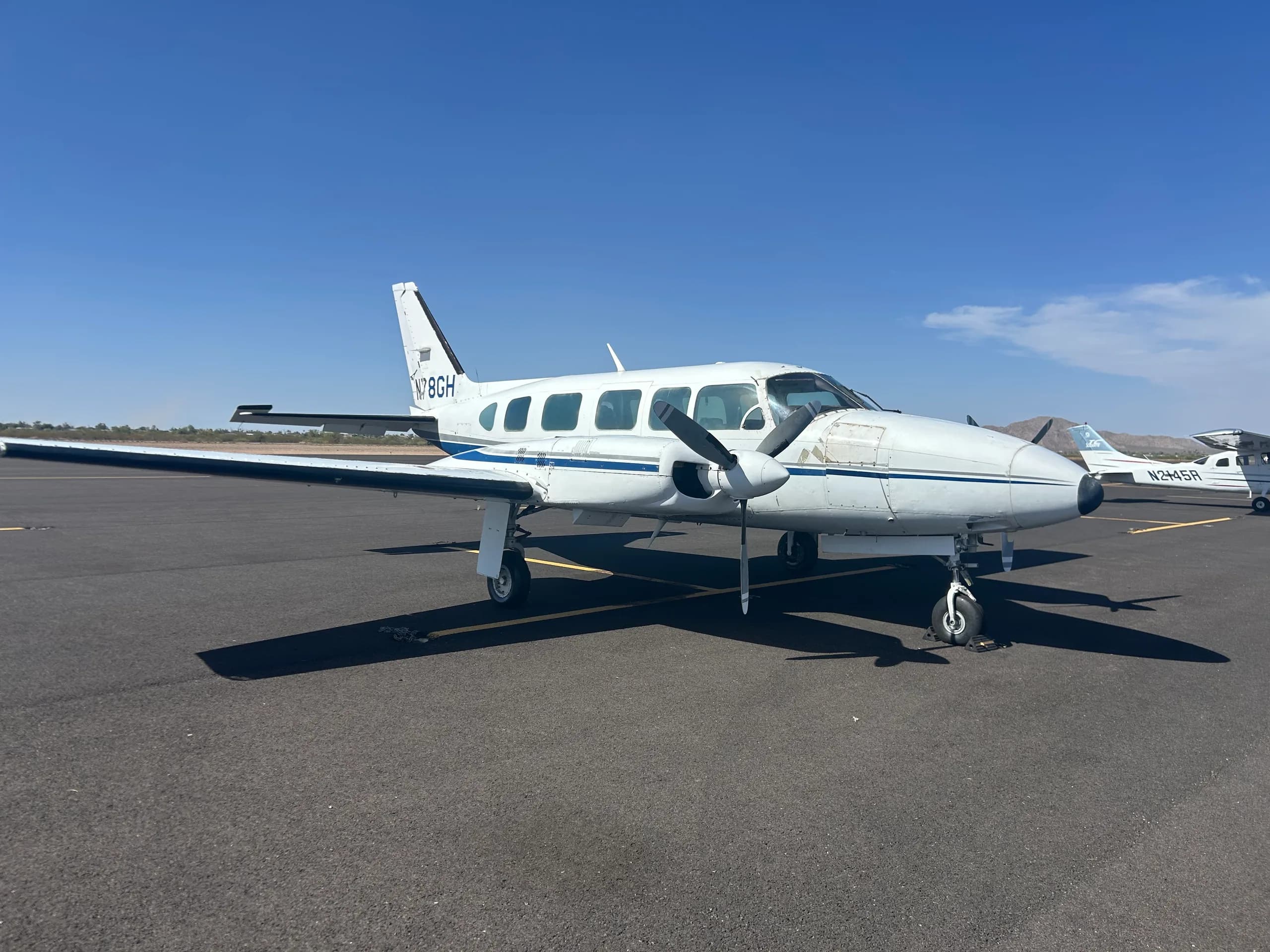A white twin-engine propeller aircraft with blue stripes, registration N78GH, parked on an asphalt tarmac under a clear blue sky.