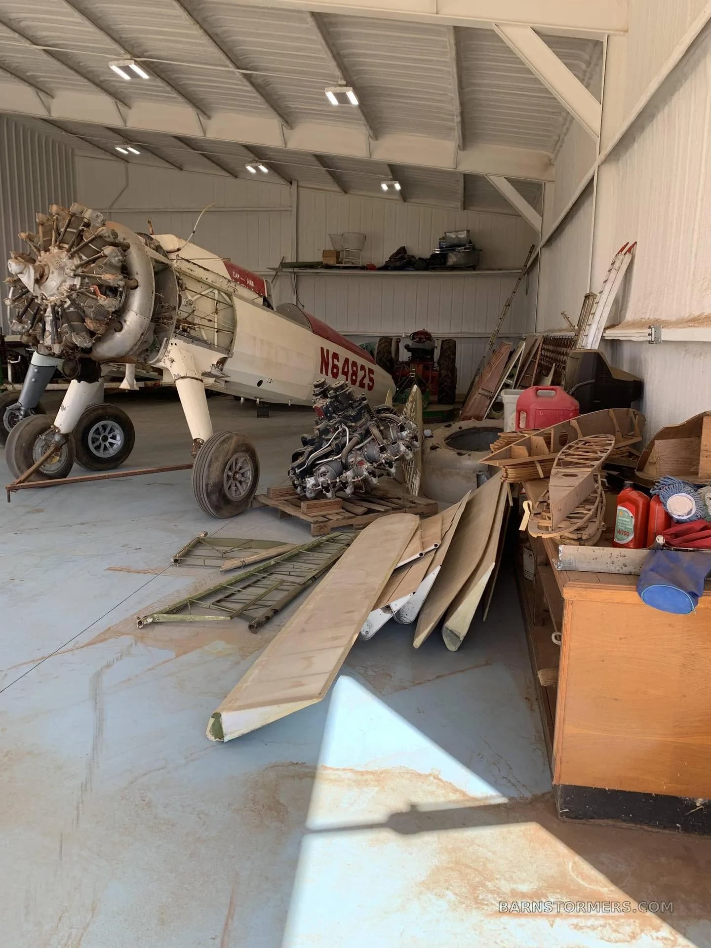 A partially disassembled white and red biplane fuselage with a radial engine, surrounded by aircraft parts and tools in a hangar.