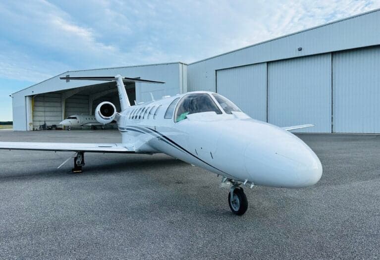 A white Cessna Citation CJ3 (N578CJ) with blue and grey stripes, parked on an asphalt tarmac in front of hangars.