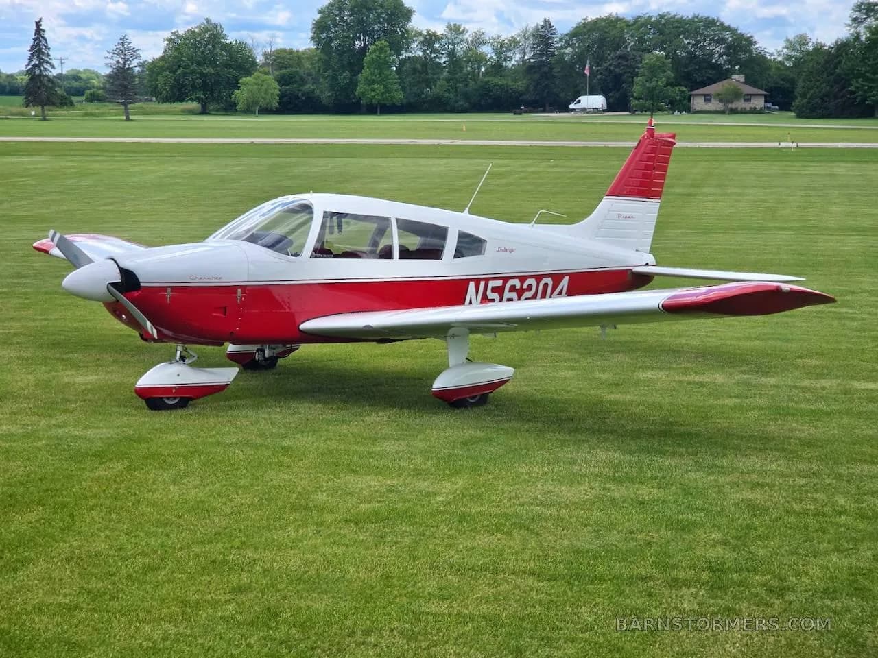 A red and white Piper Cherokee aircraft (N56204) is parked on a green grassy field under a partly cloudy sky.
