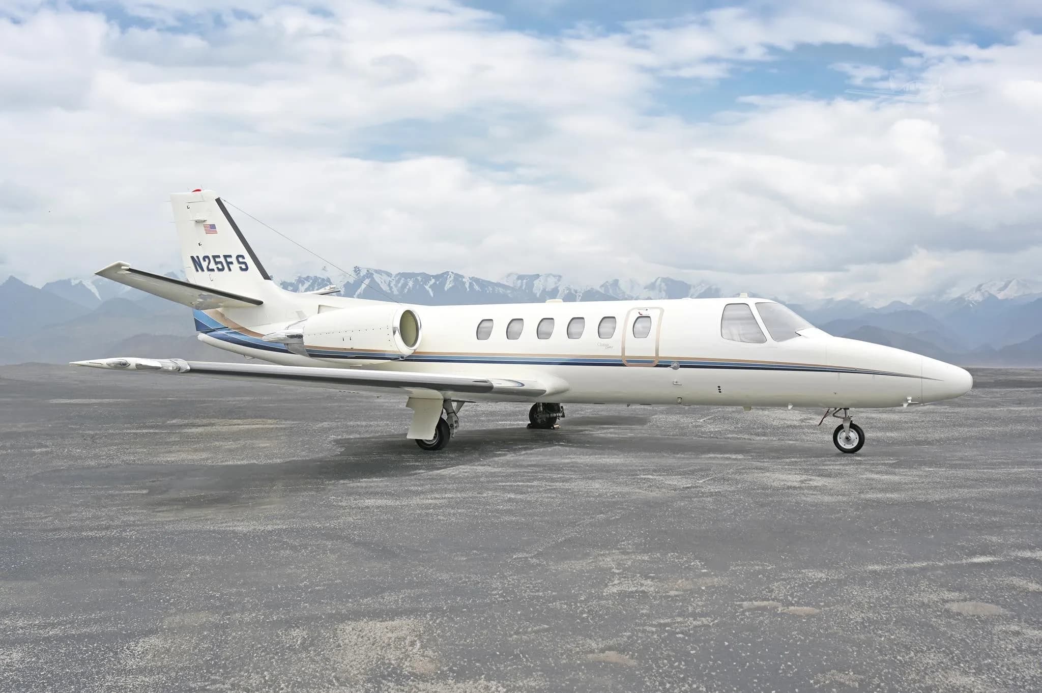 A white Cessna Citation business jet with blue and gold stripes is parked on a tarmac with mountains in the background.