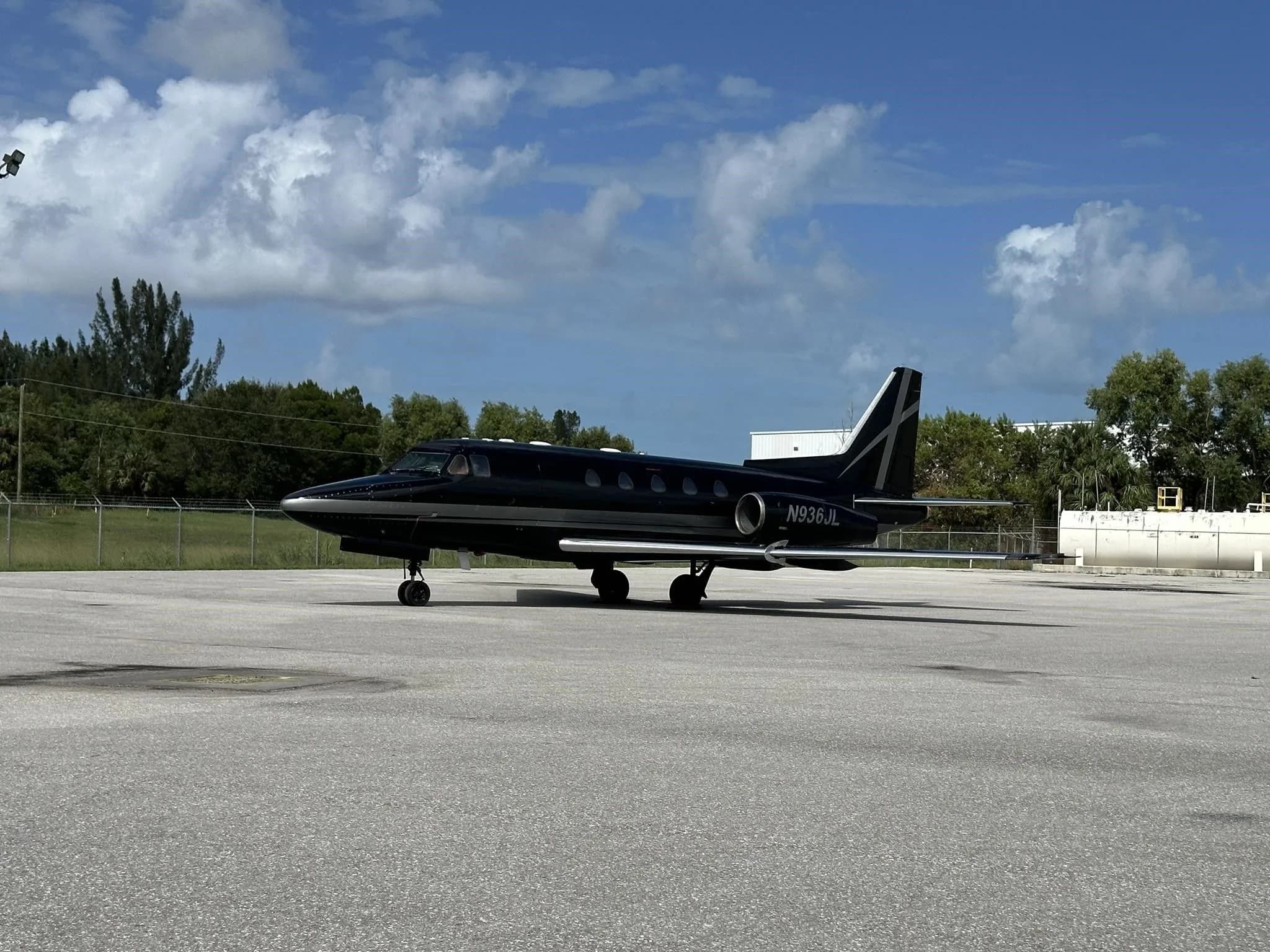 A black and silver business jet, N936JL, parked on an empty asphalt tarmac under a cloudy sky.