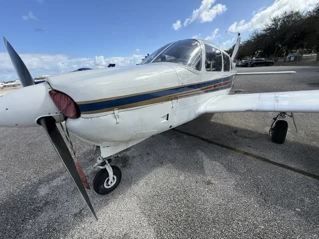 A front-side view of the white Piper Cherokee aircraft, showing its propeller, landing gear, and blue and red stripes.