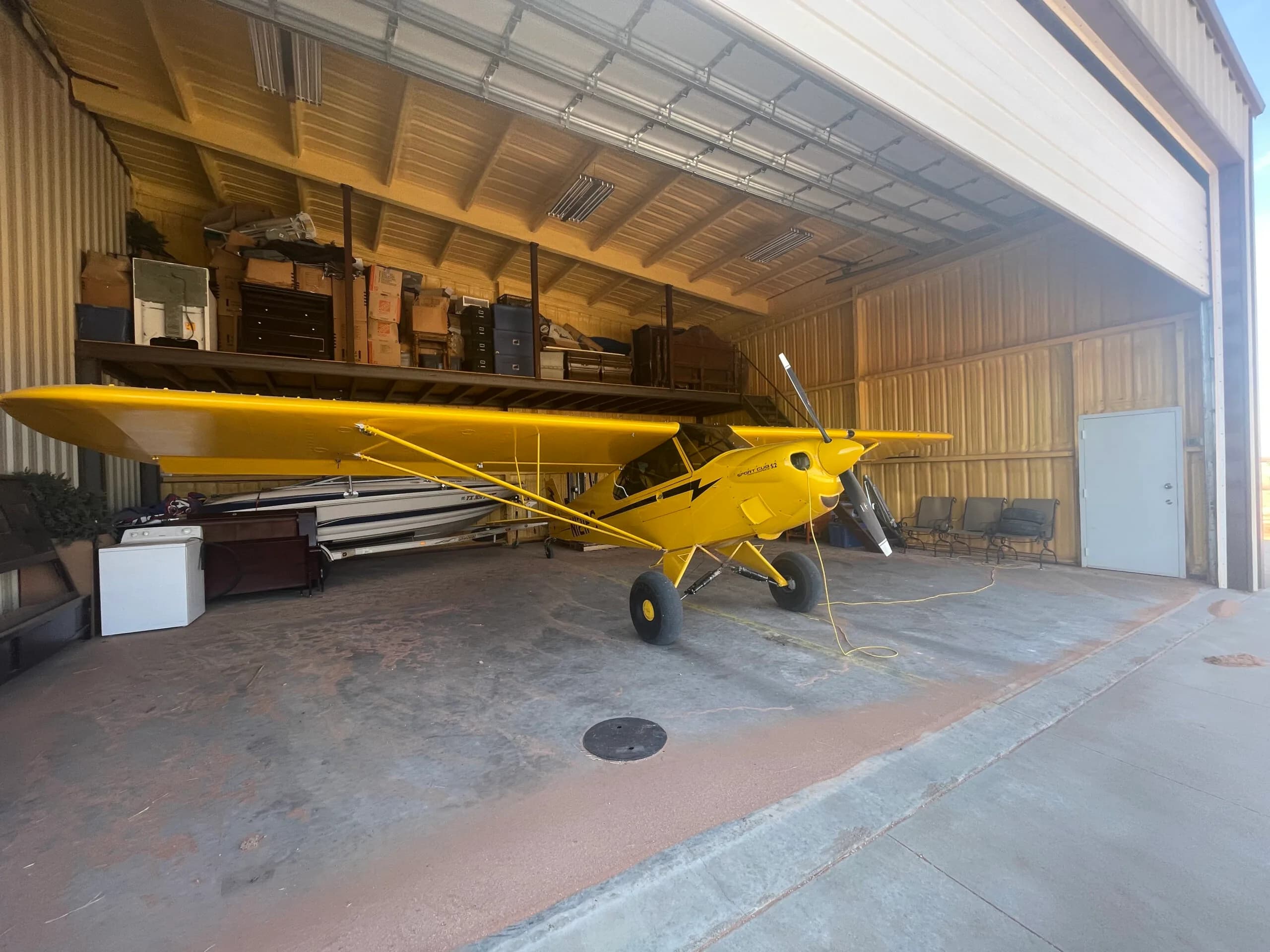 A yellow Sport Cub S2 aircraft with black stripes is parked inside a hangar, with a boat and other storage items in the background.