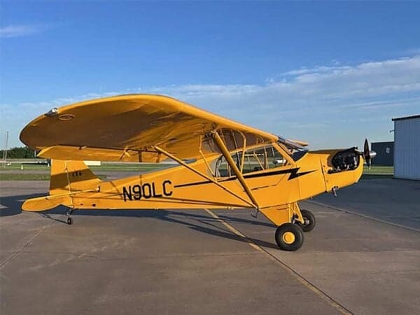 A yellow Legend Cub aircraft, tail number N90LC, parked on an airfield under a clear sky.