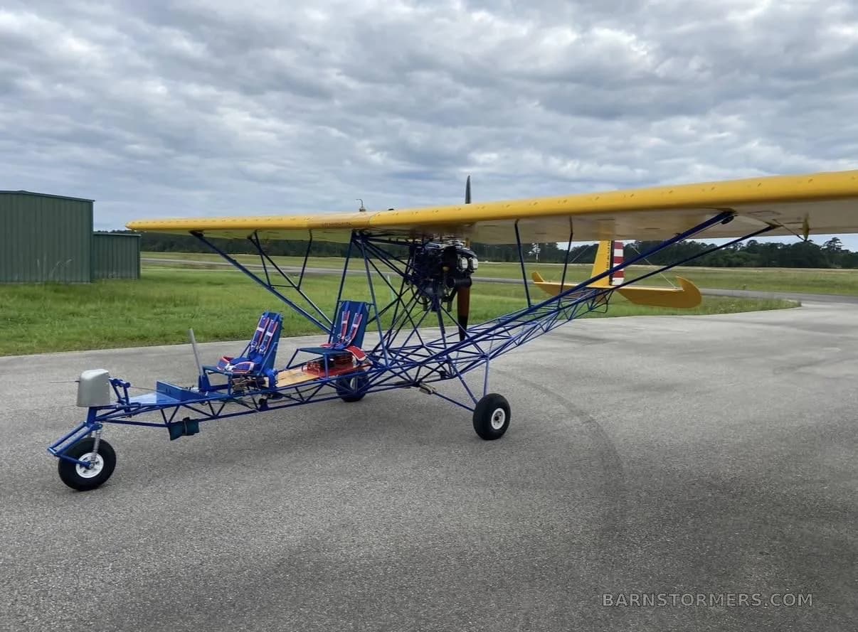A yellow-winged, blue-framed ultralight aircraft with two seats is parked on an asphalt runway under a cloudy sky.
