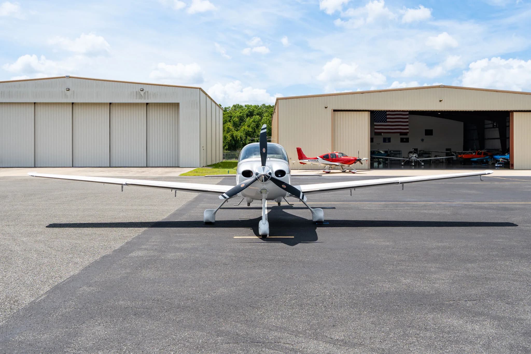 Front view of a silver Cirrus SR22 aircraft parked on an airfield with hangars in the background.