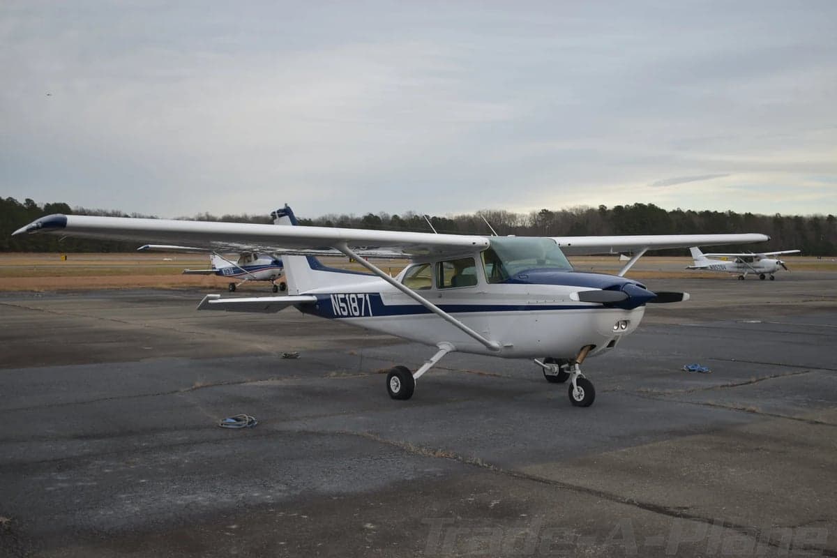A white Cessna 172 with blue and grey stripes is parked on a tarmac, viewed from the front-left with other aircraft in the distance.