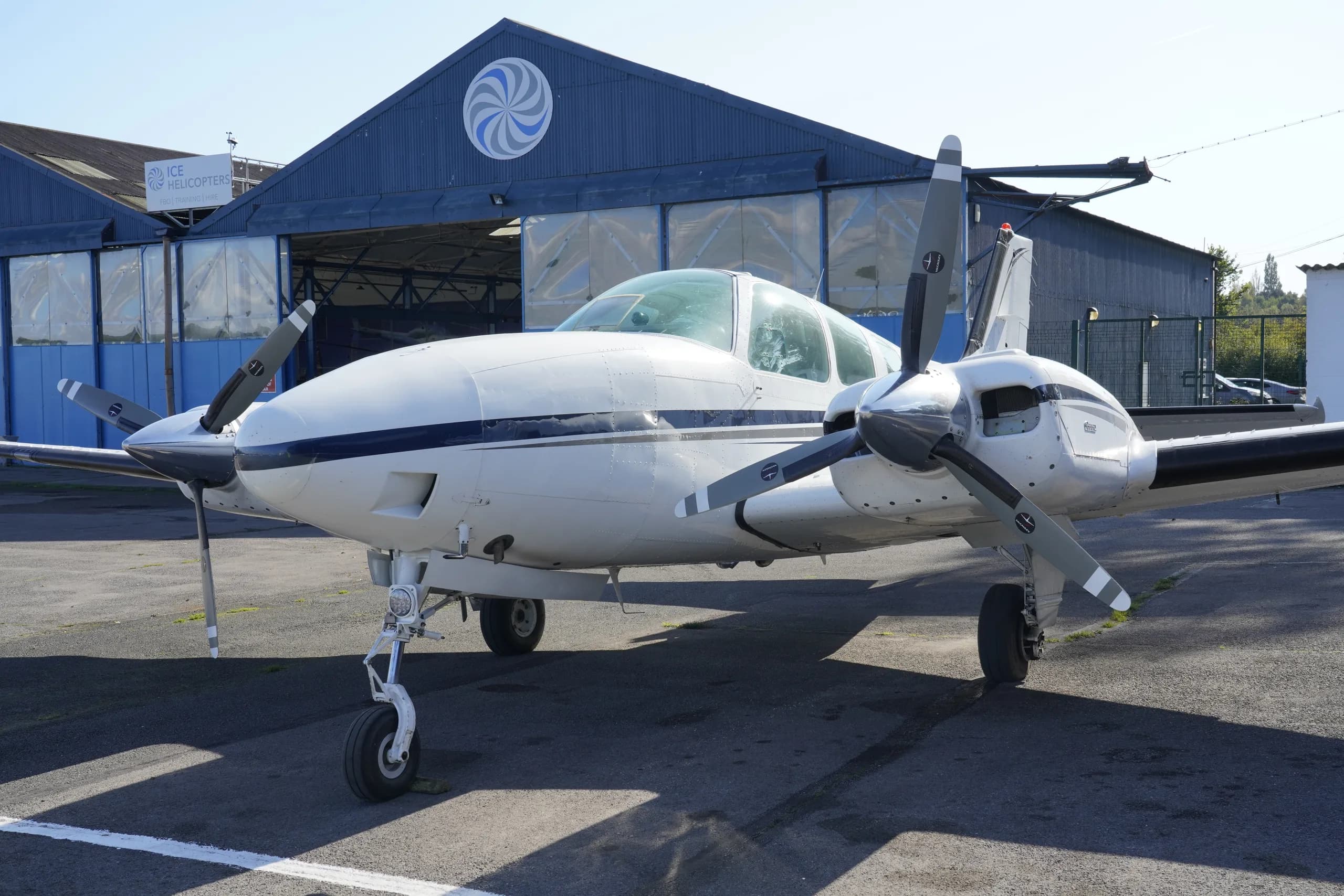 A white twin-engine propeller aircraft with blue stripes, parked on an asphalt apron in front of a blue hangar with a circular logo.