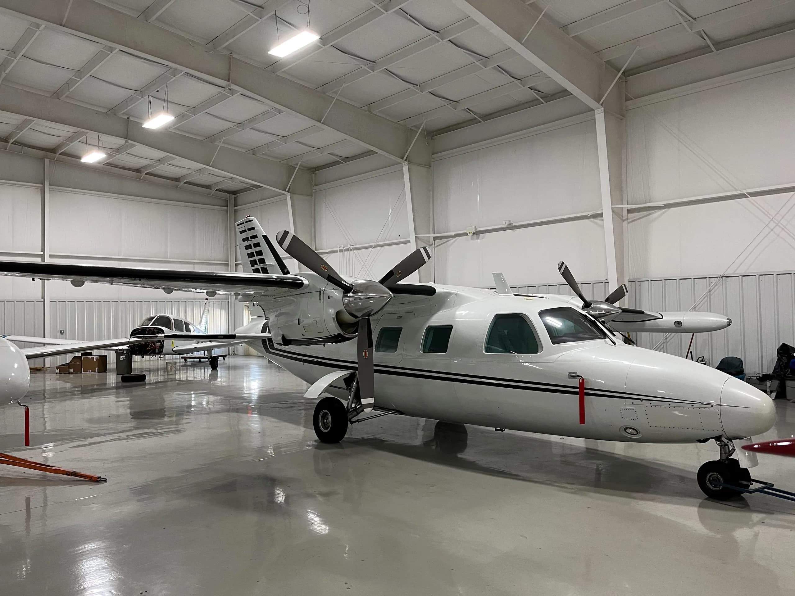 A white twin-engine turboprop aircraft with black stripes is parked in a brightly lit hangar, with another smaller aircraft visible in the background.