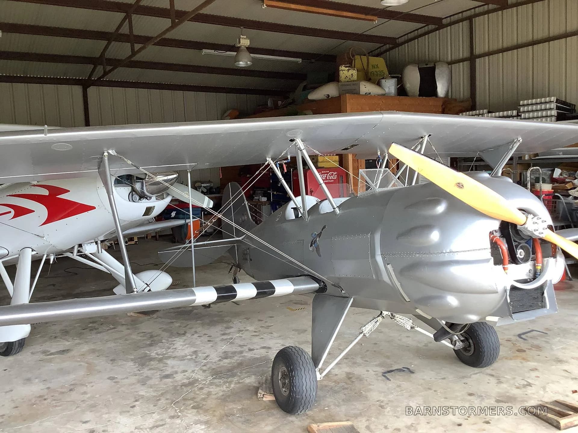 A silver biplane with a wooden propeller is parked in a hangar, with another white aircraft partially visible behind it.