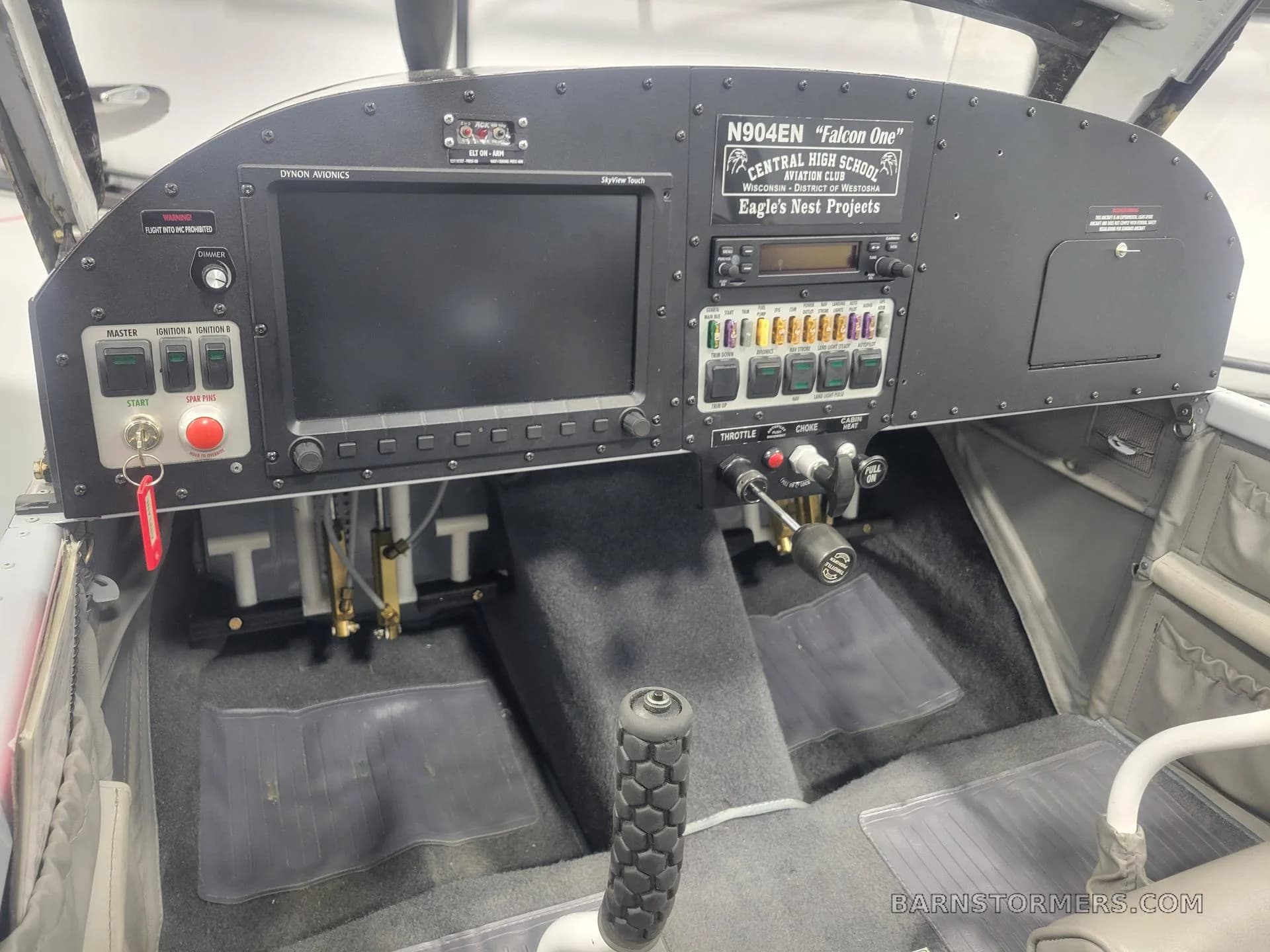 The cockpit of a single-engine aircraft, showing a Dynon avionics display, various switches, and flight controls, with a 'Falcon One' placard.
