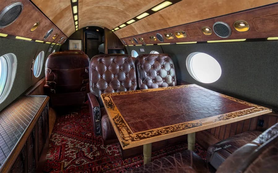 Aircraft interior with leather seats, a table, and a patterned rug.