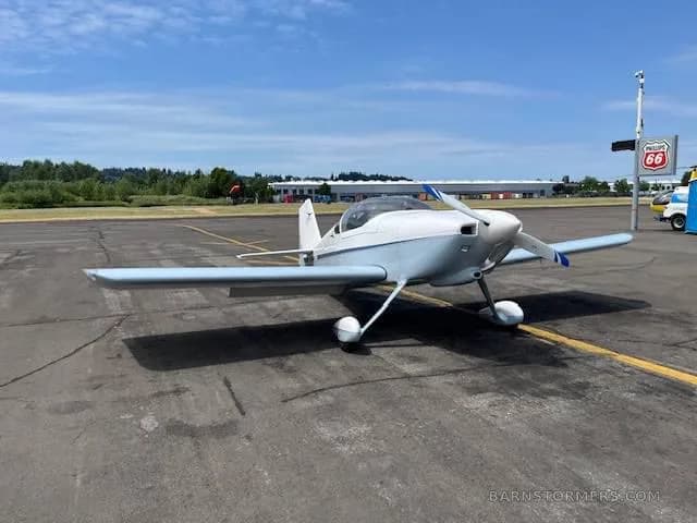 A light blue and white single-engine aircraft with a propeller, parked on an asphalt tarmac under a clear sky.