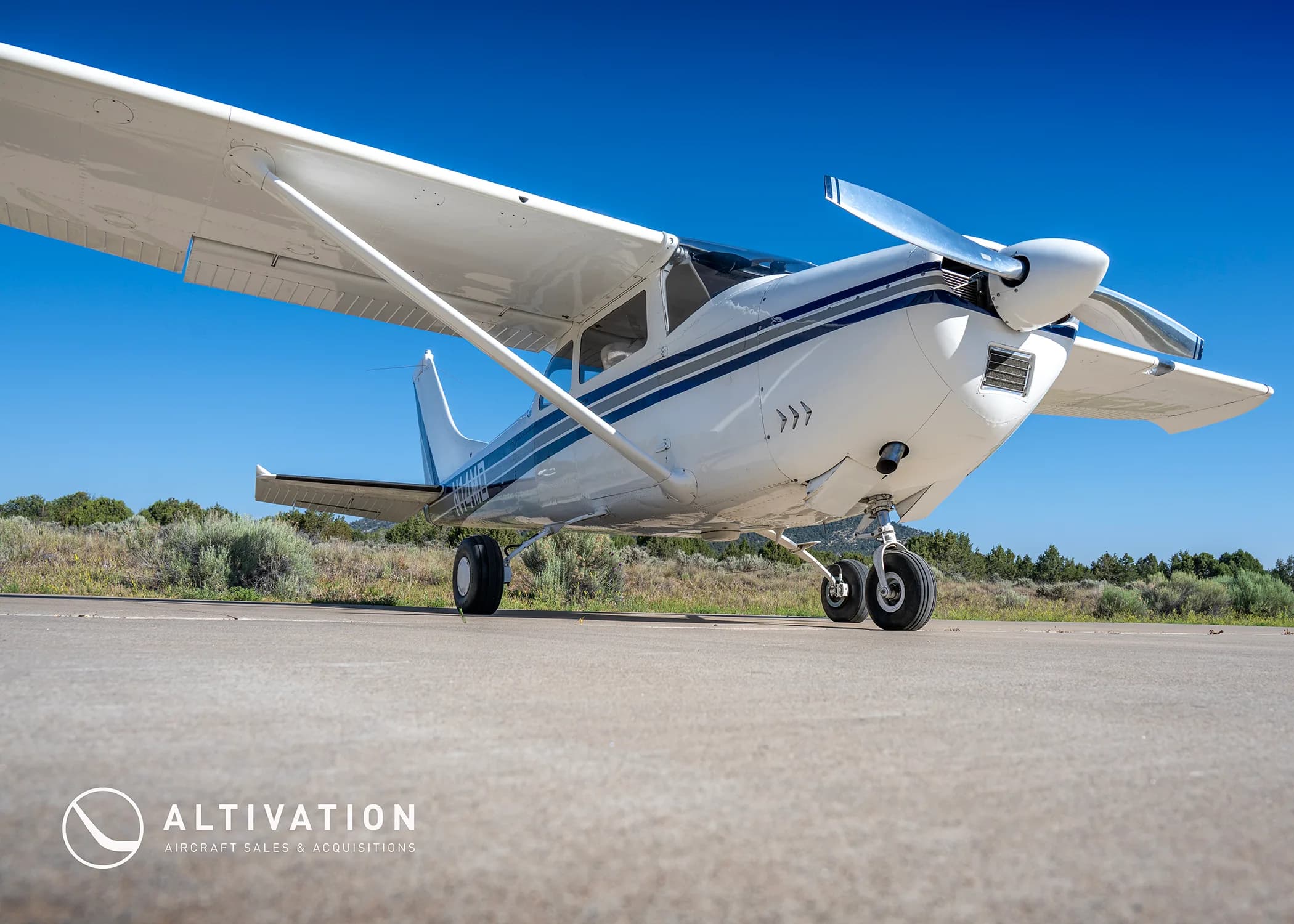 A low-angle front view of a white Cessna 182 with blue and grey stripes, registration N74MD, parked on a tarmac.