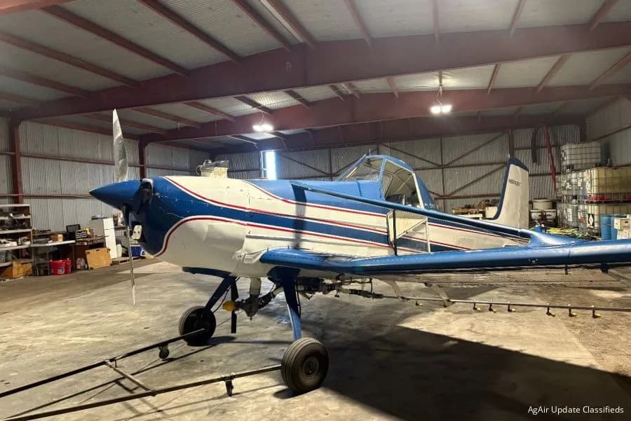 A blue, white, and red agricultural aircraft with its cockpit open, parked inside a hangar.